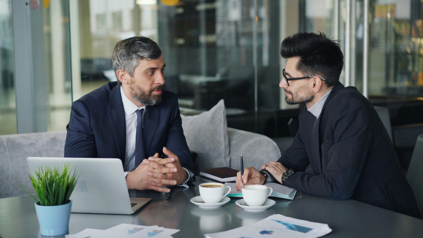 Two men in suits discussing business over coffee and a laptop in a modern office.