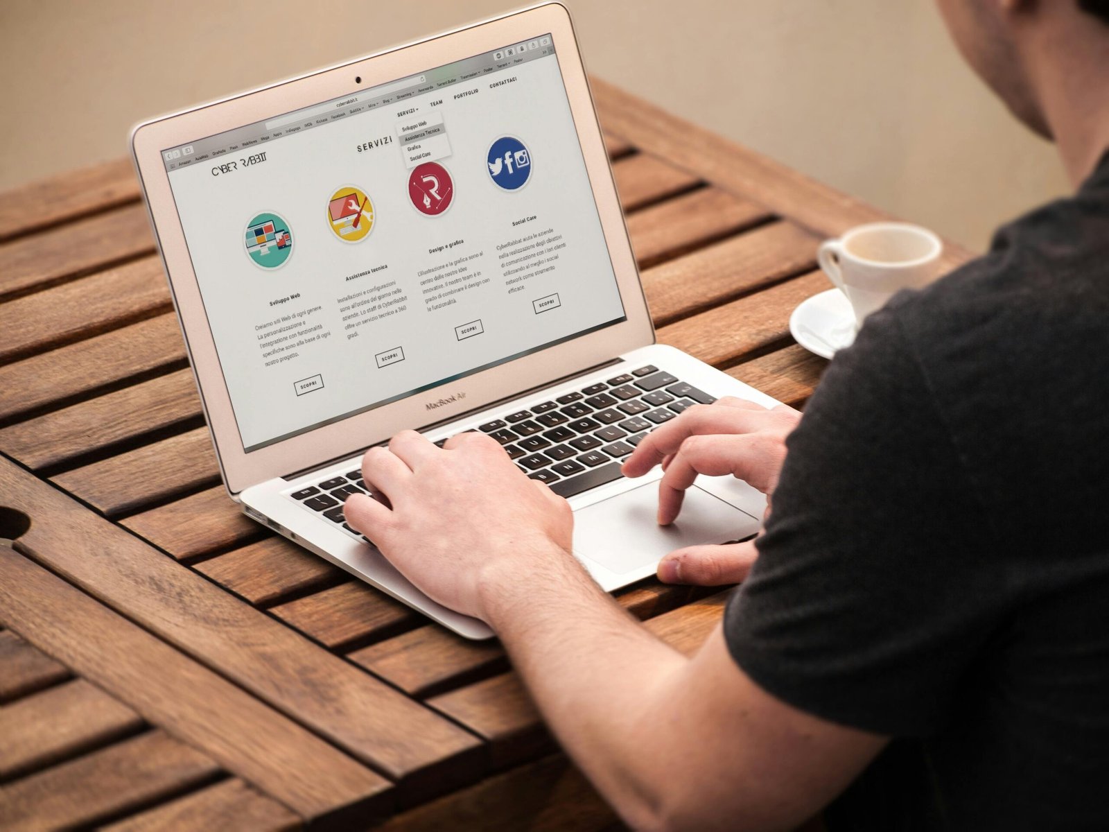 An over-the-shoulder view of a person using a MacBook Air on a wooden slatted table, displaying a "Servizi" (Services) webpage with icons for web development and social media.