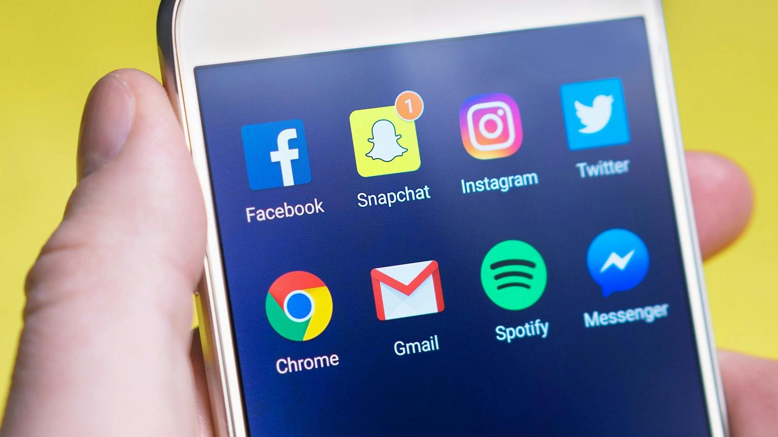 Popular Social Media and Communication Apps on Smartphone A close-up of a hand holding a smartphone showing a screen with various app icons including Facebook, Snapchat, Instagram, Twitter, Chrome, Gmail, Spotify, and Messenger.