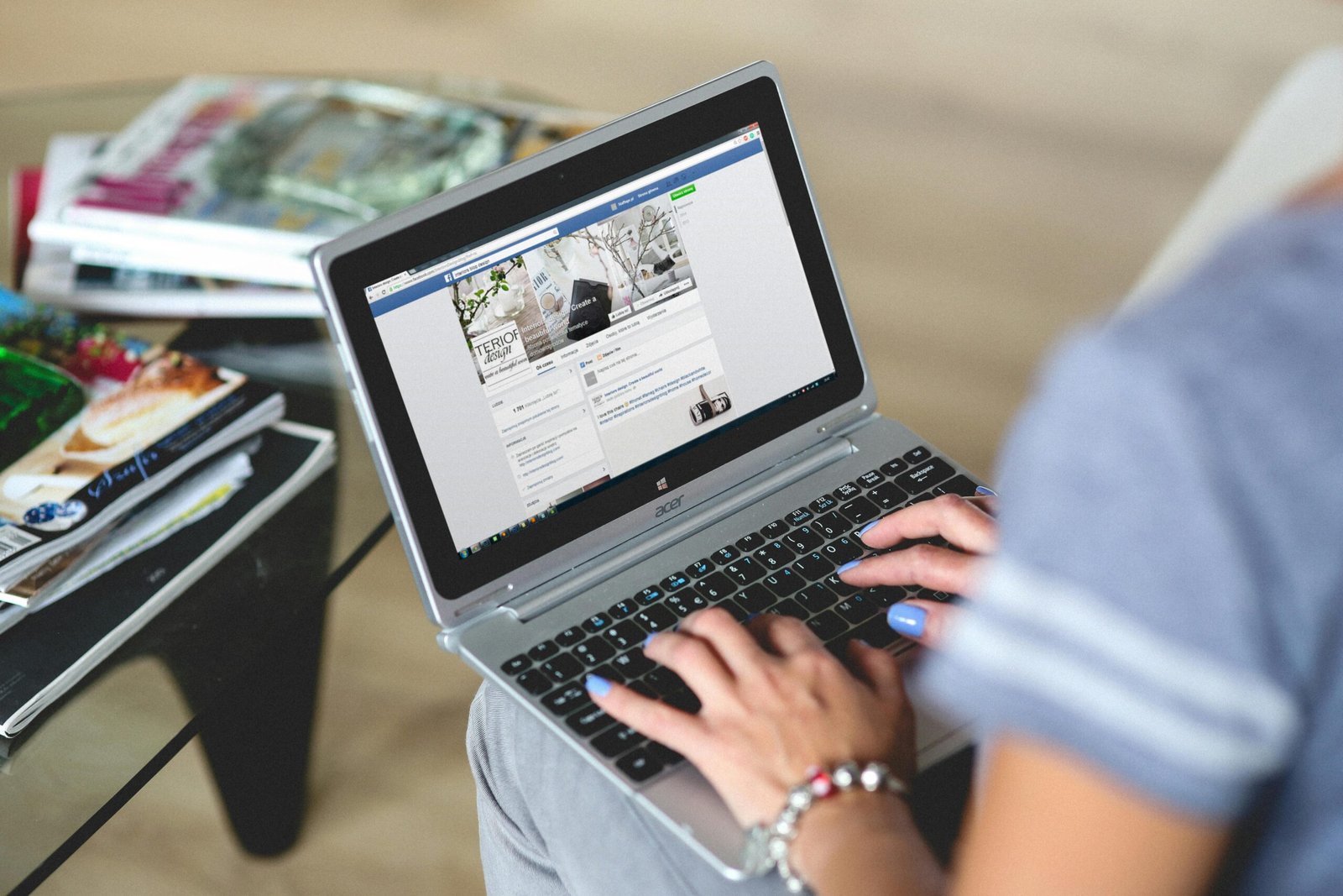 A person with blue nail polish and a bracelet typing on an Acer laptop, displaying an interior design business page on Facebook.